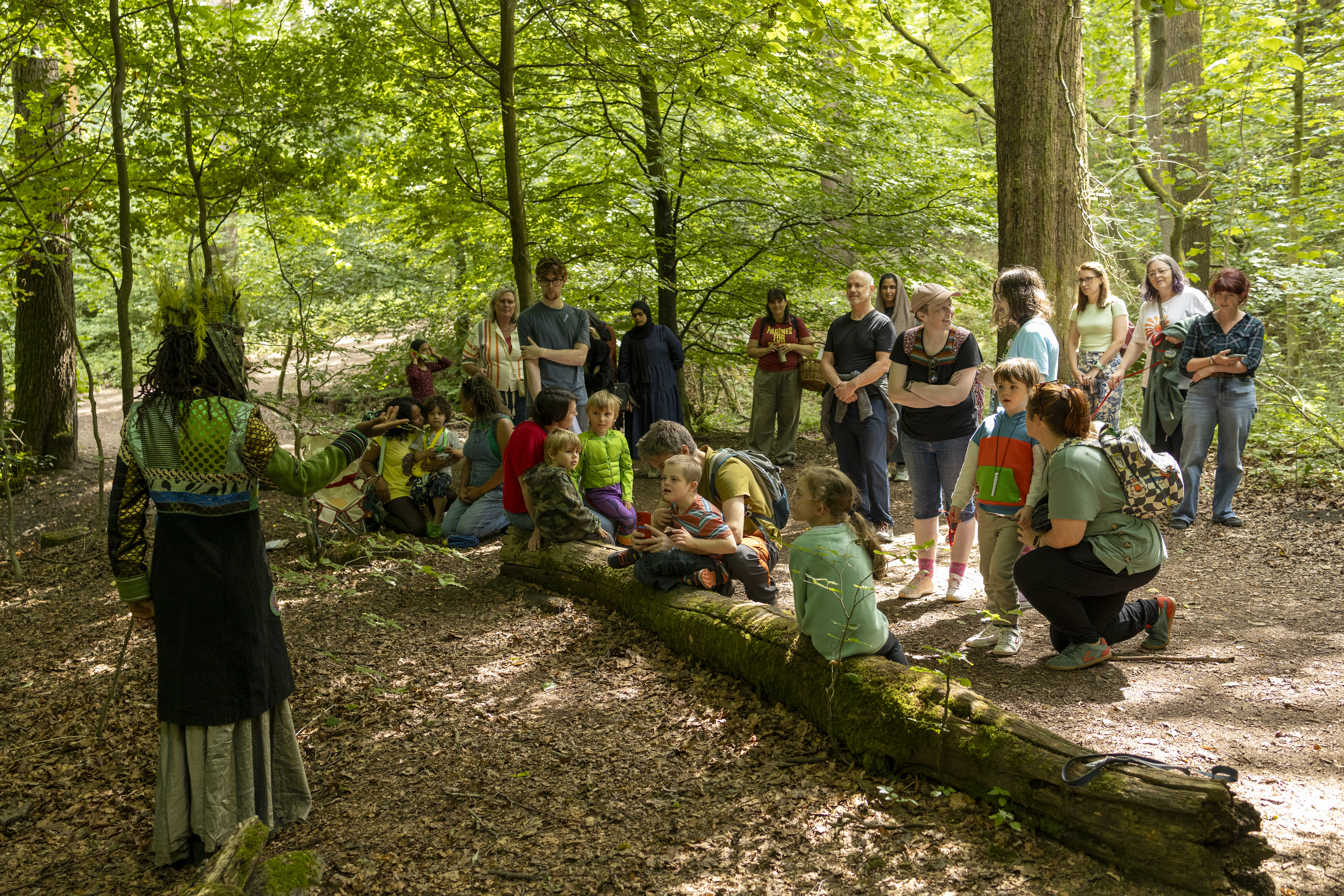 Several families and their young children in the woods gathered around a log listening to a performer