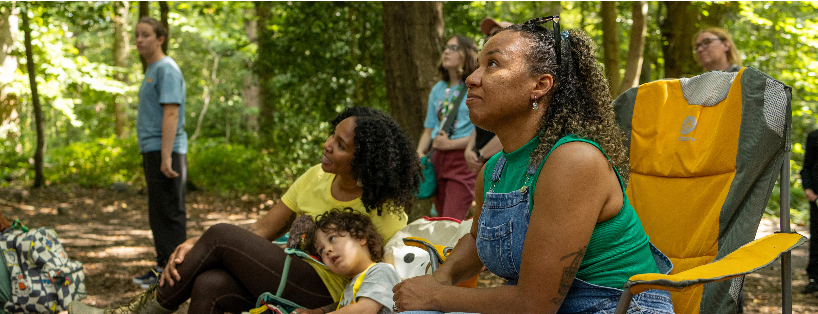A family sit down in the forest and glance upwards watching the 'A Future for All' circus show.