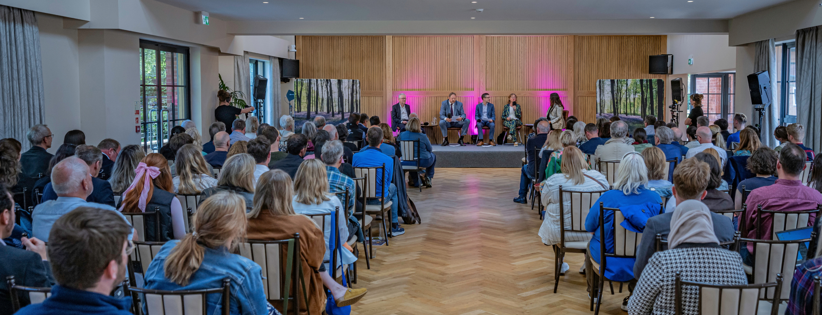A panel of five people seated on a stage, speaking to an audience seated in rows of chairs.