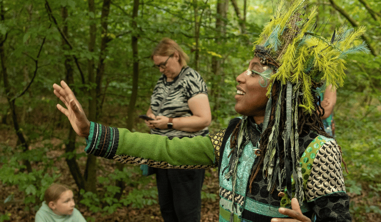 A woman wearing a botanical headdress makes a reaching gesture with her hand in a forest