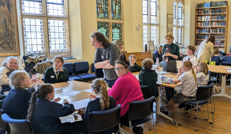 Adults and children sit at multiple tables in a room with stained glass windows and bookcases