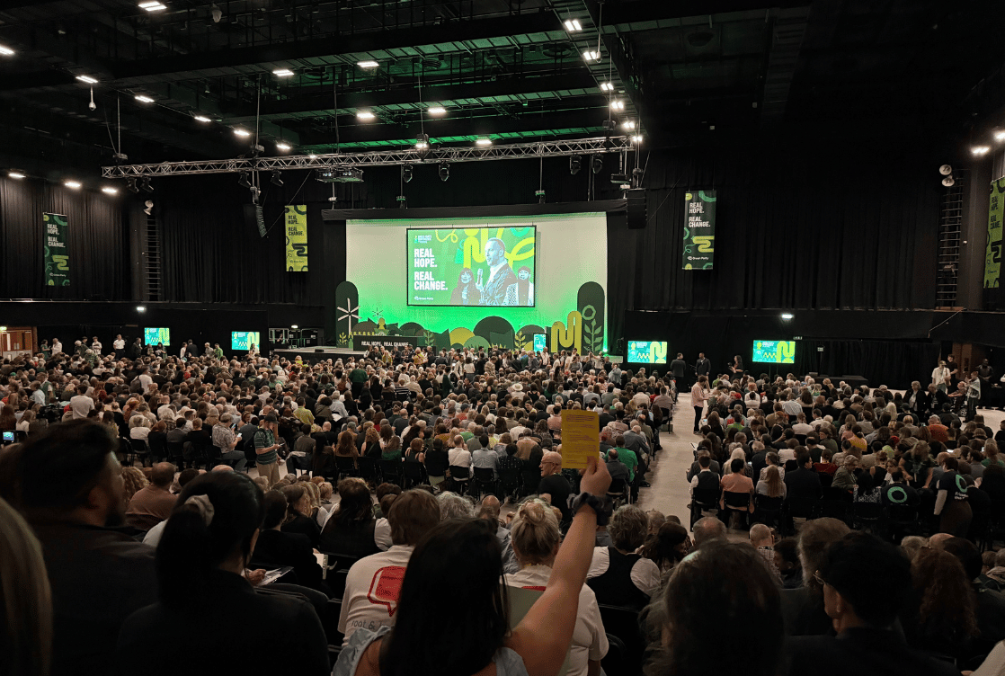 A packed conference hall faces a stage with bright green presentation graphics as attendees fill the rows.