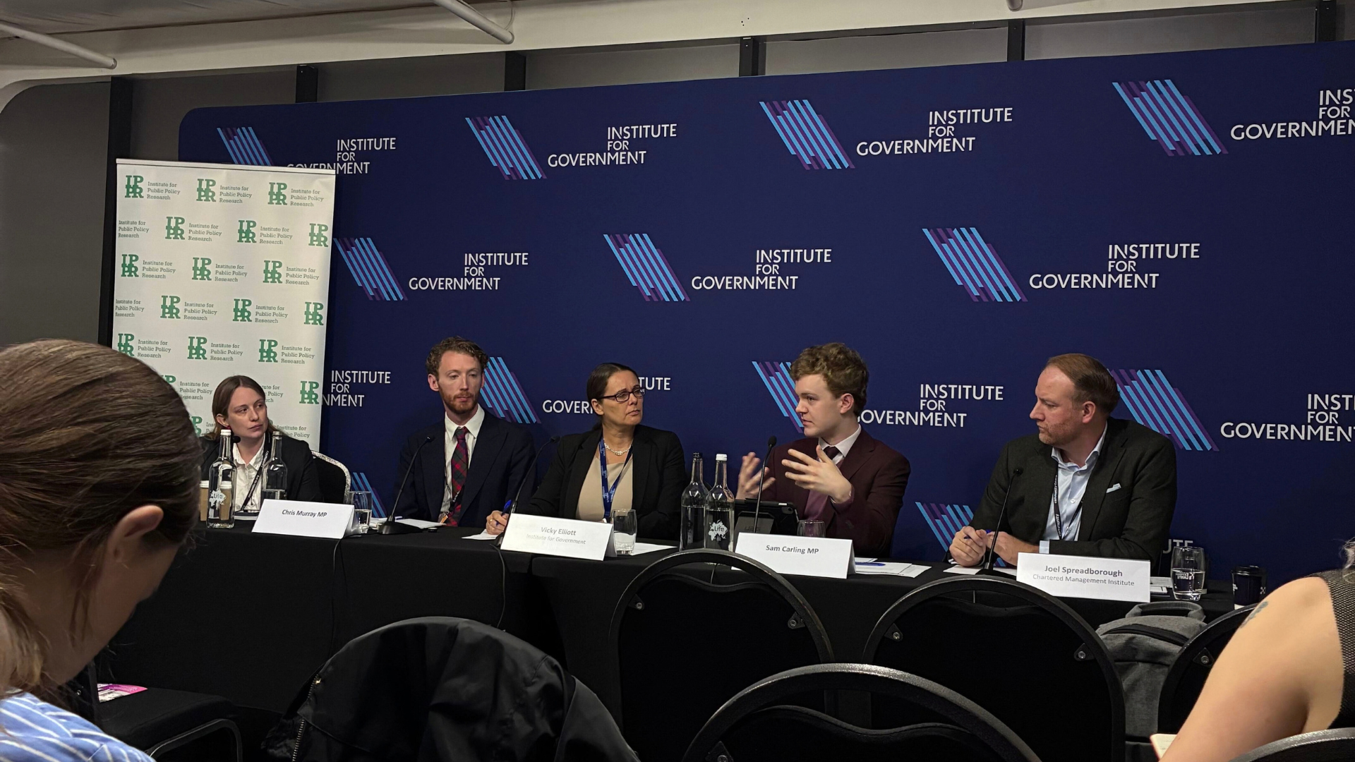 Panel of speakers seated at a conference table in front of a blue event backdrop, with microphones and name placards