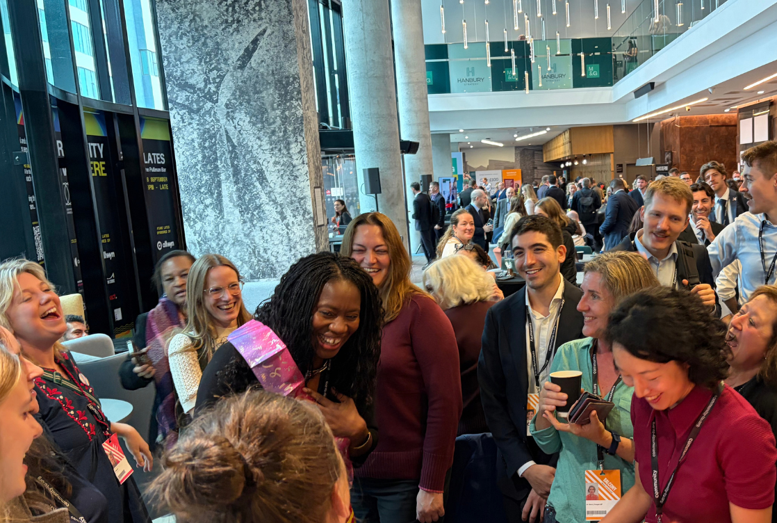 Crowd at an indoor event cheering around a birthday cake on a colorful table, with people smiling, holding drinks.