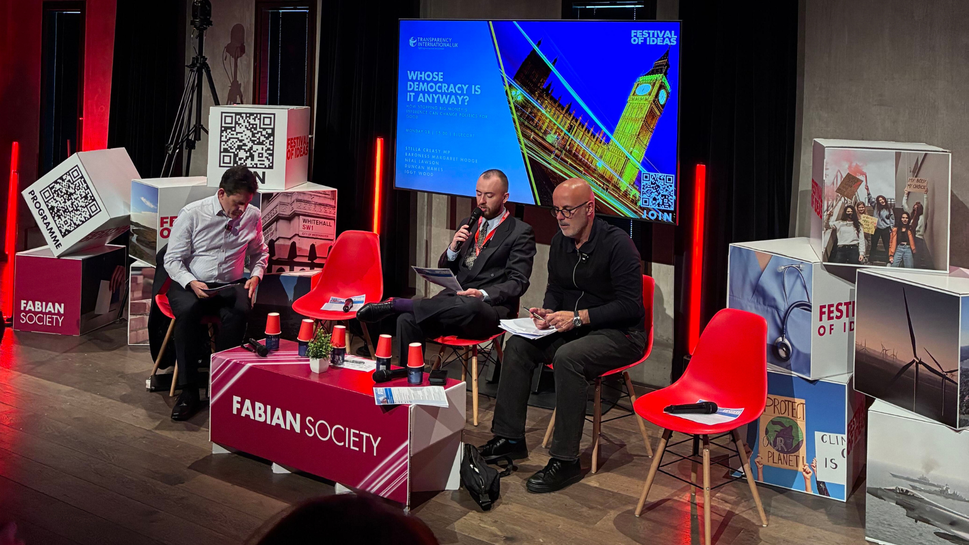 Panel discussion on a stage with four seated speakers around a table with red lighting.