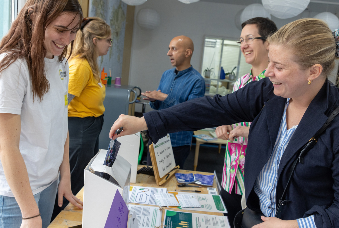 People browse a table of books and gift bags indoors; a smiling group stands and chats while one person points at items on display.