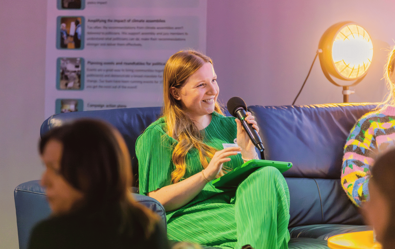 Blonde woman sits on a sofa holding a microphone in a green jumpsuit