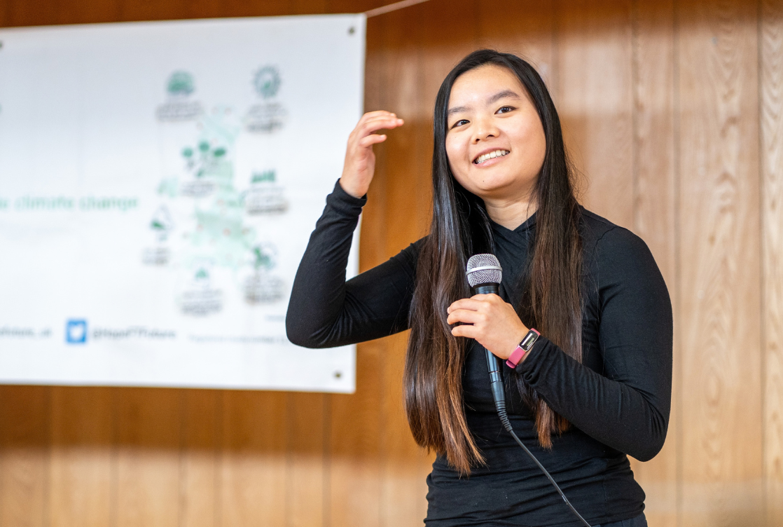 A woman with long dark hair speaking into a microphone in front of a poster about climate change.
