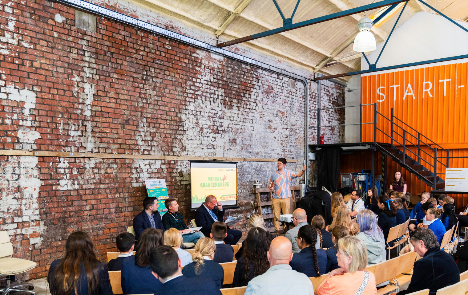 Presenter stands in front of a roll-down board speaking to an audience in an industrial warehouse